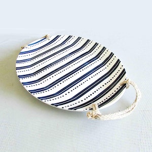 May include: Oval serving platter with a white background and navy blue stripes and dotted lines. The platter has rope handles on each side. A decorative piece for serving food or displaying items.