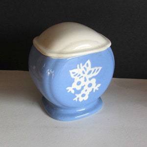 May include: A blue ceramic sugar bowl with a white lid and a white floral design. The bowl has a rounded shape and a square base.