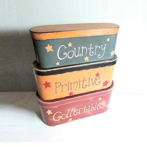 May include: Three stacked, oval-shaped wooden boxes with lids. The top box is teal with "Country" in white script and gold stars. The middle box is yellow with "Primitive" in red script and stars. The bottom box is red with "Collectibles".