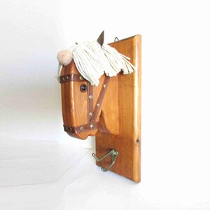 May include: Wooden horse head wall hook with a white yarn mane and a brown leather strap. The hook is attached to a wooden plaque with a metal hook underneath.