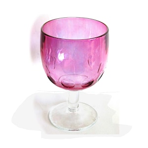 May include: A pink iridescent wine glass with a clear stem and base. The glass has a rounded bowl shape with a textured pattern near the bottom. The stem is slender, and the base is circular.