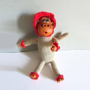 May include: A crocheted monkey toy with a red hat and red accents on the arms and legs. The monkey has a brown face and large eyes.