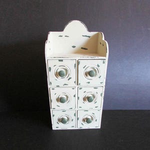 May include: A distressed white wooden cabinet with six small drawers and a decorative top. The cabinet has green accents and round, green knobs. The piece is designed for wall mounting and has a vintage, rustic aesthetic.