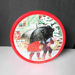 May include: A round, red-rimmed tray featuring a winter scene illustration. Two figures walk under umbrellas in the snow, carrying gifts. The artwork includes a house with lit windows and text that reads "Jenny Nyström / BUS 2009."