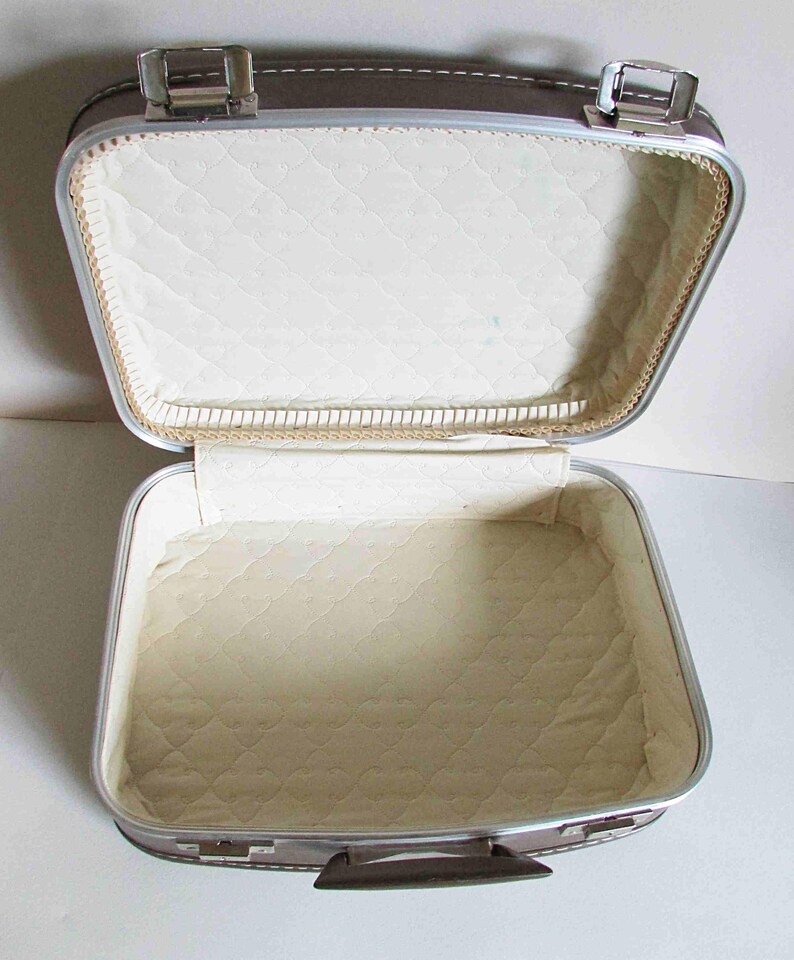 Puede incluir: Una maleta vintage plateada y marr&oacute;n con un forro acolchado blanco. La maleta est&aacute; abierta y el interior es visible.