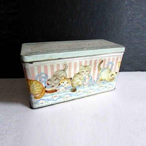 May include: A rectangular metal tin with a lid, featuring a pastel pink and white striped pattern. The tin is decorated with illustrations of playful kittens and blue ribbon. The lid has the word "Coupons" in cursive.