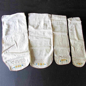 May include: Four white linen bags with embroidered lettering. The bags are labeled "Knives", "Forks", "Salad", and "Teaspoons".