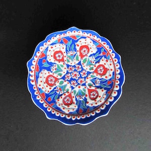 May include: A decorative ceramic plate with a scalloped edge. The plate features a vibrant floral design in red, white, and blue, with green accents. The intricate pattern is centered on a dark background.