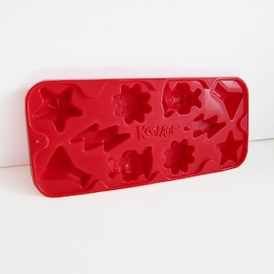 May include: A red silicone ice cube tray with various festive shapes, including stars, flowers, and lightning bolts. The tray is marked with the "Kool-Aid" brand name.
