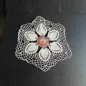 May include: A handmade, white lace doily with a floral design. The center of the doily is a reddish-orange color, surrounded by six white, leaf-shaped patterns. The doily is placed on a dark surface.