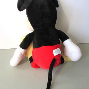 May include: A plush Mickey Mouse toy, viewed from the back. The toy features a black head and ears, a red bottom, yellow torso, white gloves, and a long black tail. The toy is sitting on a light gray surface.
