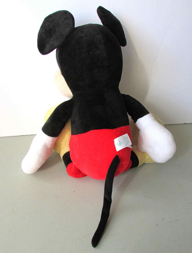 May include: A plush Mickey Mouse toy, viewed from the back. The toy features a black head and ears, a red bottom, yellow torso, white gloves, and a long black tail. The toy is sitting on a light gray surface.