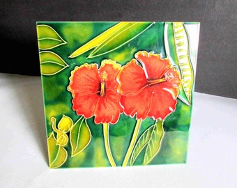 May include: A ceramic tile with a green background and two red hibiscus flowers. The flowers have yellow centers and are surrounded by green leaves.