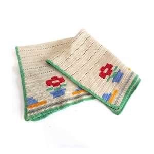 May include: A cream-colored, crocheted blanket with a green border. The blanket features a repeating pattern of red flowers in blue pots, with yellow and gray accents. The blanket is folded, showing the pattern on both sides.