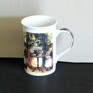 May include: White ceramic mug featuring a detailed illustration of two rabbits in a kitchen setting. One rabbit wears a hat and apron, the other a dress. The handle has a brown and tan pattern.