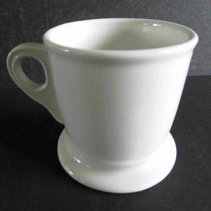 Op de afbeelding: Een witte keramische mok met een handvat en een brede, licht uitlopende rand. De mok staat op een kleine, afgeronde basis. De algehele vorm is een klassiek, eenvoudig ontwerp.
