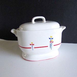 May include: A white ceramic tureen with a matching lid and two side handles. The exterior features a horizontal red line accented with small, stylized floral motifs in blue, yellow, and red. The vessel rests on an oval base with a red trim.