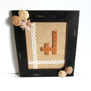 May include: A black framed picture with a burlap background and white lace trim. The words "MOTHER" and "YOU" are spelled out using wooden letter tiles. The picture is decorated with brown fabric flowers.