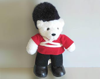 May include: A plush teddy bear dressed as a British Royal Guard. The bear has a white face and paws, a black furry hat, a red jacket with white straps, black pants, and black boots. The toy is standing upright.