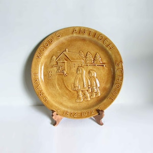 May include: A brown ceramic plate with a raised design of a schoolhouse and two children walking. The plate is inscribed with the text "Grant Wood's Antioch School 1872-1972".