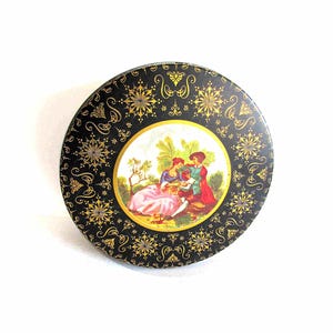 May include: Round black tin with a gold floral and snowflake pattern. The center features a painted scene of a couple in period clothing. The tin has a gold rim and is set against a white background.