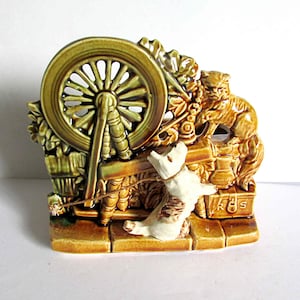 May include: A ceramic planter shaped like a spinning wheel with a cat and a dog figurine. The wheel is green and brown, the cat is brown, and the dog is white. The planter is marked "R.S."
