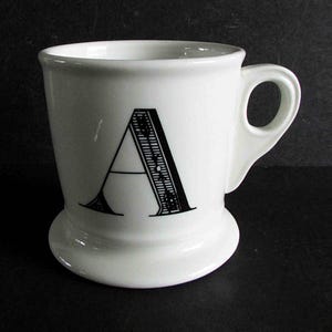 Op de afbeelding: Een witte keramische mok met de zwarte letter "A". De mok heeft een afgeronde basis en een handvat. De letter "A" is versierd met lijnen en patronen. Geschikt voor warme dranken.