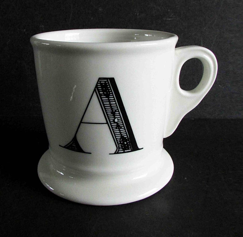Op de afbeelding: Een witte keramische mok met de zwarte letter "A". De mok heeft een afgeronde basis en een handvat. De letter "A" is versierd met lijnen en patronen. Geschikt voor warme dranken.