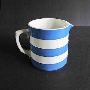 May include: A ceramic pitcher featuring a horizontal striped pattern in bright blue and white. The vessel has a smooth, cylindrical body, a curved handle on the side, and a small pouring spout at the rim. The interior of the pitcher is white.