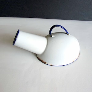 May include: A white enamel bedpan with a blue handle and rim. The vintage medical item has a cylindrical spout and a rounded base. The rim shows signs of age and wear.