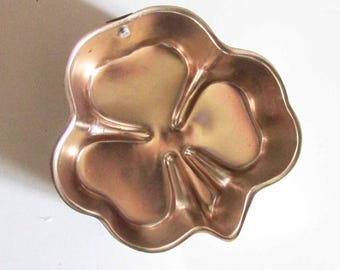 Puede incluir: Un molde para hornear en forma de flor, de color cobre. El molde tiene un diseño de tres lóbulos, con bordes redondeados y un pequeño agujero en la parte superior para colgarlo. El interior tiene una superficie lisa y reflectante.