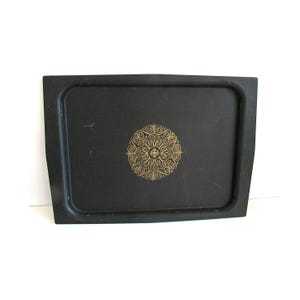 May include: A rectangular black tray with a gold-colored circular design in the center. The design is an intricate pattern of swirls and curves. The tray has slightly curved edges and a smooth surface.