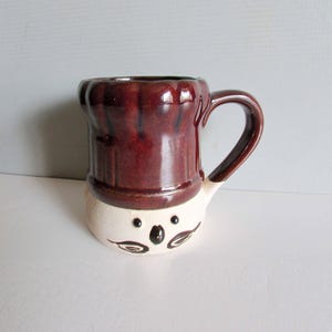 May include: A ceramic mug with a chef-hat design. The top portion is a deep burgundy color, while the bottom is off-white with a cartoon face. The mug has a curved handle and is suitable for hot or cold beverages.