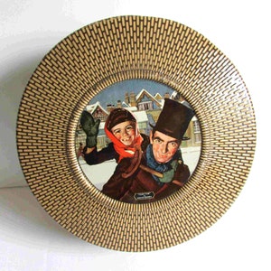 May include: Round decorative plate with a woven pattern border in shades of tan and brown. The center features a vintage illustration of two people in winter clothing, playing in the snow. The plate has a glossy finish.