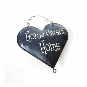 May include: A heart-shaped, dark blue metal sign with the words "Home Sweet Home" in white script. The sign has a wire hanger at the top and a small loop at the bottom. The sign is designed for home decor.