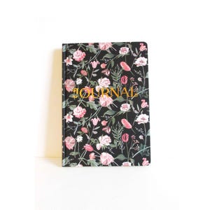 May include: A floral journal with a black cover featuring pink and white flowers and green foliage. The word "JOURNAL" is printed in gold lettering across the front. The journal is rectangular and appears to be new.