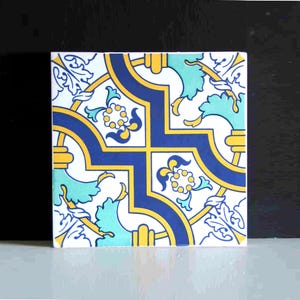 May include: Square ceramic tile with a repeating pattern in blue, yellow, and teal on a white background. The design features floral and geometric elements, creating a decorative and symmetrical look. The tile is suitable for home decor.