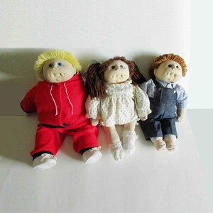 May include: Three soft-sculpture dolls with yarn hair and round faces. The left doll has yellow hair and a red tracksuit. The center doll has brown pigtails and a white floral dress with white socks. The right doll has brown hair and denim overalls.