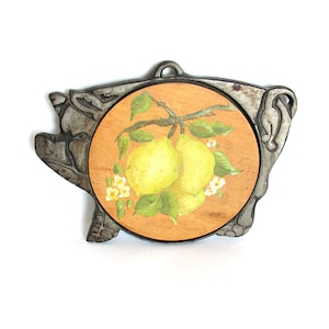 May include: A decorative trivet with a painted lemon design. The round wooden center features yellow lemons, green leaves, and white flowers on a branch. The trivet is framed by a decorative metal border with a pig-like shape.