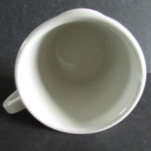 Op de afbeelding: Bovenaanzicht van een witte keramische mok met een handvat. De mok heeft een eenvoudig, afgerond ontwerp en een gladde, glanzende afwerking. De binnenkant van de mok is zichtbaar en toont een schoon, lichtgekleurd oppervlak. De mok staat tegen een donkere achtergrond.