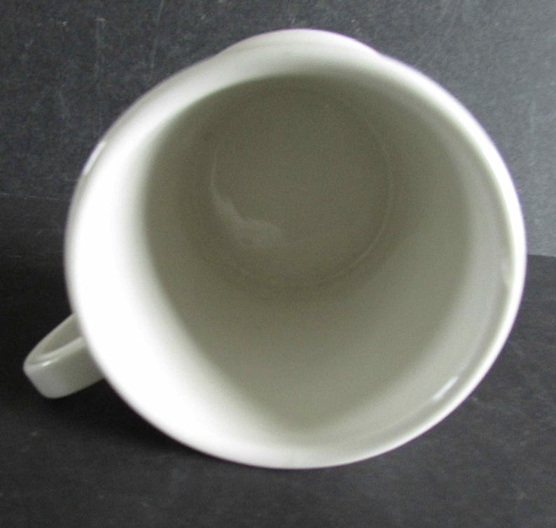Op de afbeelding: Bovenaanzicht van een witte keramische mok met een handvat. De mok heeft een eenvoudig, afgerond ontwerp en een gladde, glanzende afwerking. De binnenkant van de mok is zichtbaar en toont een schoon, lichtgekleurd oppervlak. De mok staat tegen een donkere achtergrond.