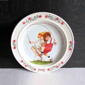May include: A white ceramic plate featuring a vintage illustration of two children jumping rope. The plate has a floral border with the word "JUNE" and text that reads "A certified plate in the original and only Sarah Stilwell Weber Calendar Collection."