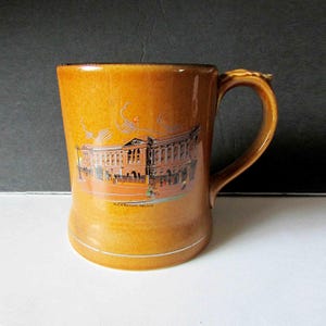May include: A tan ceramic mug with a detailed illustration of Buckingham Palace. The mug has a curved handle and a slightly textured surface. The words "Buckingham Palace" are printed below the image.