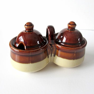 May include: A brown and cream ceramic double condiment dish with lids and a handle. The dish has a glazed finish.