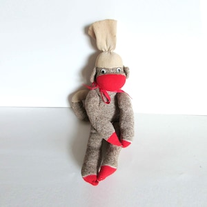May include: A brown sock monkey with a beige sock hat and red felt accents. The monkey has googly eyes and a red felt mouth.