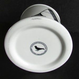 Op de afbeelding: Witte keramische mok met een zwart en grijs geometrisch ontwerp. De bodem van de mok heeft een zwart vogel logo en de tekst "Dishwasher & Microwave Safe Made in China Fabriqu&eacute; en Chine."