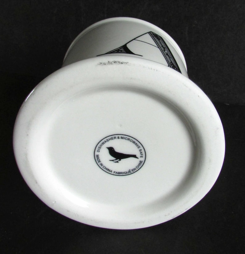 Op de afbeelding: Witte keramische mok met een zwart en grijs geometrisch ontwerp. De bodem van de mok heeft een zwart vogel logo en de tekst "Dishwasher & Microwave Safe Made in China Fabriqu&eacute; en Chine."