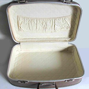 Puede incluir: Una maleta vintage marr&oacute;n con un forro acolchado blanco y un peque&ntilde;o bolsillo dentro de la tapa. La maleta tiene un marco de metal plateado y un asa marr&oacute;n.