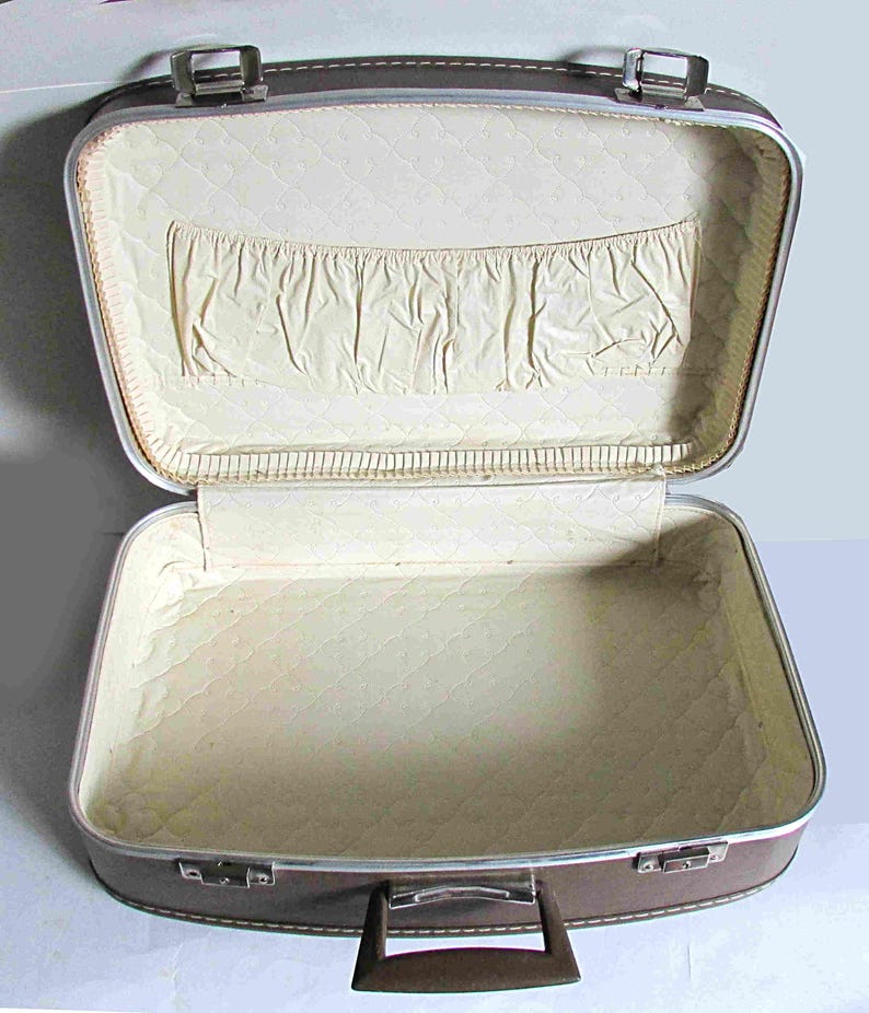 Puede incluir: Una maleta vintage marr&oacute;n con un forro acolchado blanco y un peque&ntilde;o bolsillo dentro de la tapa. La maleta tiene un marco de metal plateado y un asa marr&oacute;n.