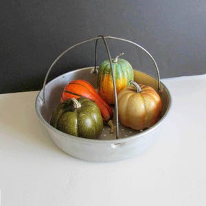 May include: A metal serving bowl with a handle holds a collection of colorful gourds. The gourds are in shades of orange, green, and yellow, and are arranged inside the bowl. The bowl has a perforated bottom and a silver handle.